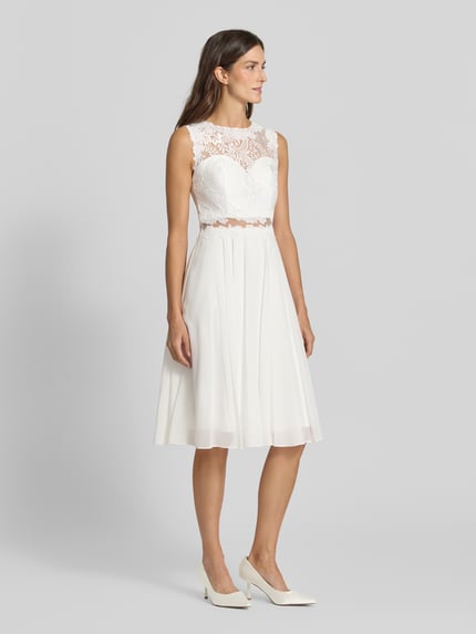 Frauenmodel trägt Cocktailkleid mit Spitze und Rundhalsausschnitt von Mascara aus weißem Chiffon in A-Linie, Midikleid, Ganzkörperansicht von vorne.