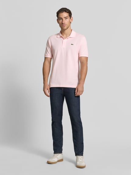 Männermodel trägt Regular Fit Poloshirt aus reiner Baumwolle von Lacoste in Hellrosa mit Kragen, kombiniert mit dunkler Jeans, Ganzkörperansicht von vorne, Herren Poloshirt von Lacoste.