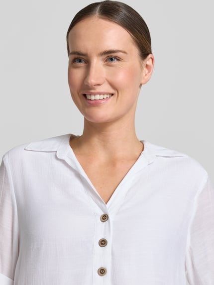 Vrouwenmodel draagt Blusenshirt met Umlegekragen van Apricot in witte viscosemix, recht model, platte kraag, V-hals, knoopsluiting, 1/2-mouwen, ruglengte maat S 82 cm, mouwlengte maat S 28 cm. Nahaufnahme bovenlichaam.