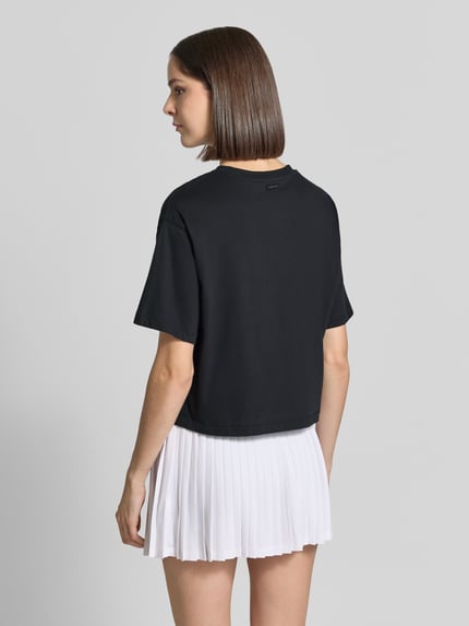 Frauenmodel trägt Relaxed Fit T-Shirt aus Baumwoll-Mix von Fabletics in Schwarz, kurzer Schnitt, kombiniert mit weißem Faltenrock, Rückenansicht.
