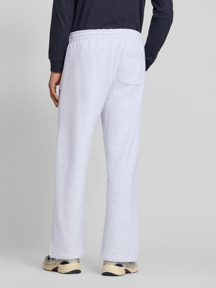 Męski model nosi spodnie dresowe Sweatpants mit elastischem Bund und Label-Print od Carhartt Work In Progress, bawełna, krój wide leg, model 'Pond Corps', elastyczny pas, szwy pionowe, nadruk z logo, długość wewnętrzna nogawki w rozmiarze M 72 cm, jasnoszary kolor, gładka faktura, widok całej sylwetki z tyłu.