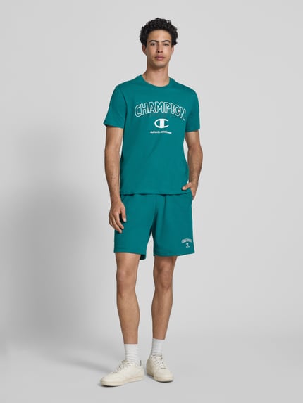 Männermodel trägt Sweatshorts mit Logo und elastischem Bund von CHAMPION, Regular Fit, Baumwoll-Mix, türkis, Ganzkörperansicht von vorne.