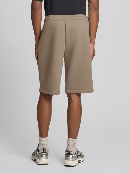 Männermodel trägt Bermudas mit Reißverschlusstaschen von EA7 Emporio Armani aus Material-Mix mit Viskose-Anteil, Regular-Fit, elastischer Bund, Farbe Beige, Ganzkörperansicht Rückansicht.