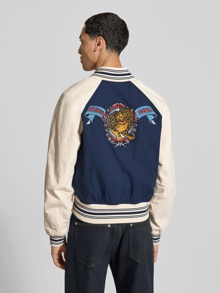 Männermodel trägt Regular Fit Collegejacke aus reiner Baumwolle von Kenzo in Blau und Beige, auffällige Rückenstickerei, Rückenansicht, Herren Collegejacke, durchgehende Knopfleiste, Eingrifftaschen, Label-Stitching, Rückenlänge 60 cm, Schulterbreite 45 cm, Ärmellänge 64 cm.