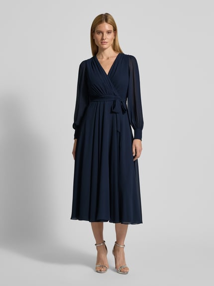 Frauenmodel trägt Regular Fit Midikleid mit Taillenband von Christian Berg Cocktail aus navyfarbenem, semitransparentem Chiffon mit blickdichtem Unterrock, V-Ausschnitt und Wickel-Optik, Falten-Textur, langärmelig; Ganzkörperansicht von vorne.