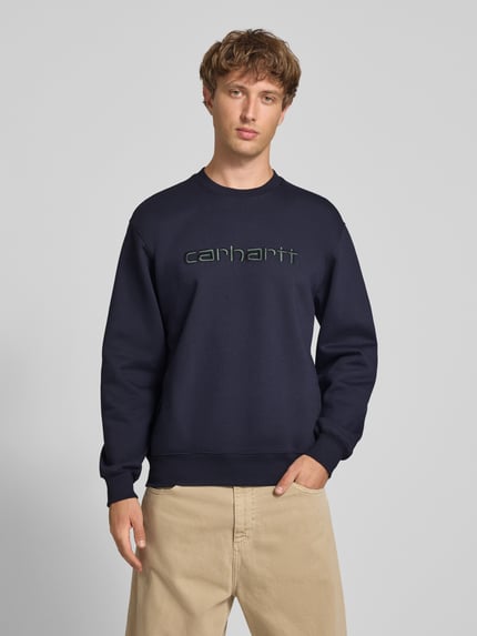 Männermodel trägt Regular Fit Sweatshirt aus Baumwoll-Mix mit Logo-Stickerei von Carhartt Work In Progress, dunkelblau, Ganzkörperansicht von vorne. Oversized Passform, gerippte Abschlüsse, langärmelig, Rundhalsausschnitt.