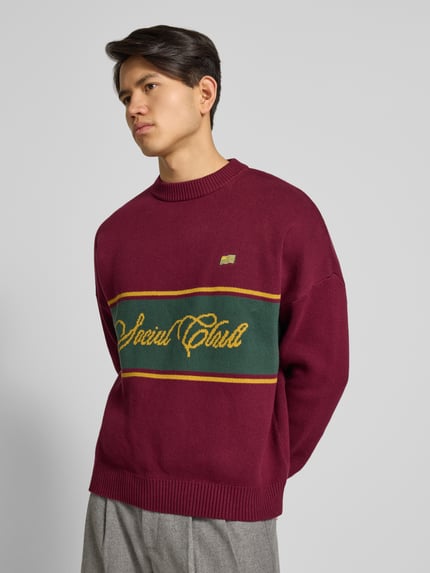 Männermodel trägt Strickpullover mit Rundhalsausschnitt von PROHIBITED in Bordeaux mit grün-gelbem Kontraststreifen, Relaxed-Fit, Reine Baumwolle, Ganzkörperansicht von vorne.