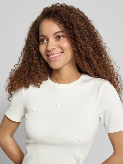 Vrouwenmodel draagt Slim Fit T-Shirt aus Baumwoll-Mix Modell 'FEMKE' van Only in effen wit met strikapplicaties en fijne ribstructuur, korte mouw, ronde hals, detailopname bovenlichaam.