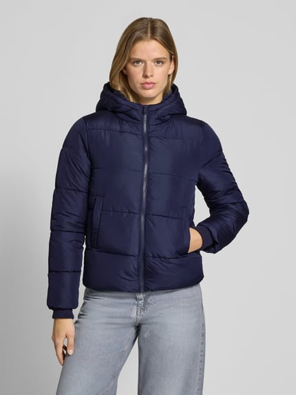 Frauenmodel trägt dunkelblaue Steppjacke mit Kapuze von PIECES, dezente Steppstruktur, kombiniert mit hellblauer Jeans, Ganzkörperansicht von vorne.