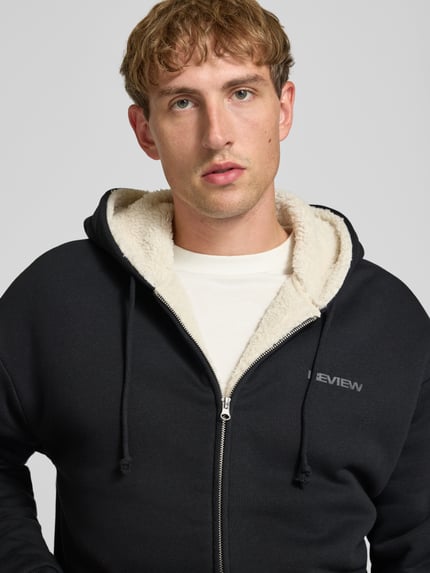 Männermodel trägt Regular Fit Sweatjacke aus Baumwoll-Mix mit Teddyfutter von REVIEW, Logo-Detail, Nahaufnahme Oberkörper.