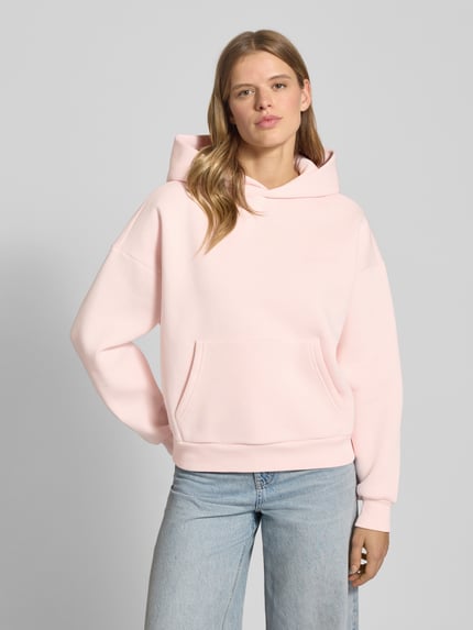 Frauenmodel trägt Oversized Hoodie mit Kapuze von Review in zartem Rosa, Baumwoll-Mix, Label-Print, Känguru-Tasche, gerippte Abschlüsse, Ganzkörperansicht von vorne.