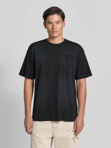 Männermodel trägt schwarzes T-Shirt mit Label-Stitching von Diesel aus reiner Baumwolle, gerade geschnitten, Brusttasche, Ganzkörperansicht von vorne.