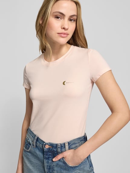Vrouwenmodel draagt T-Shirt met Label-Print van ARMANI EXCHANGE in lichtroze, slim fit, katoen-elastaan, close-up bovenlichaam.