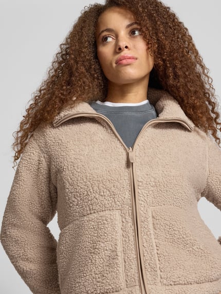 Frauenmodel trägt Teddyjacke mit hohem Stehkragen von ONLY in Beige, Fleece-Struktur, Reißverschluss, aufgesetzte Taschen, Nahaufnahme Oberkörper.