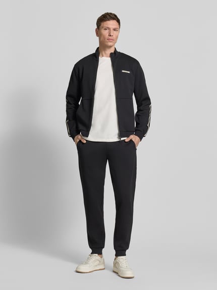 Männermodel trägt Sweatjacke mit Stehkragen und Zweiwege-Reißverschluss von EA7 Emporio Armani in Schwarz, Regular Fit, Ganzkörperansicht von vorne.