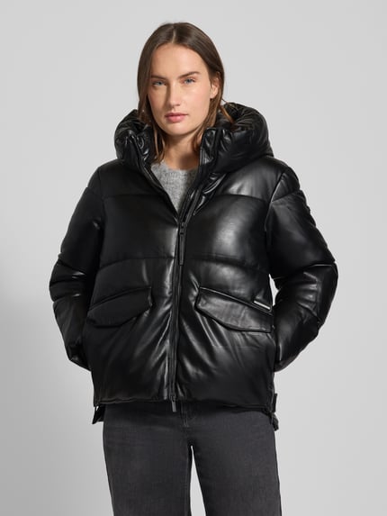 Frauenmodel trägt schwarze Steppjacke mit Kapuze Modell 'AURORA' von khujo, Regular Fit, Reißverschluss, Pattentaschen, Label-Detail, Ganzkörperansicht von vorne. Rückenlänge 65 cm, Schulterbreite 44 cm, Brustweite 50 cm, Ärmellänge 60 cm bei Größe S. Glatte, glänzende Textur mit großflächigen Steppnähten.