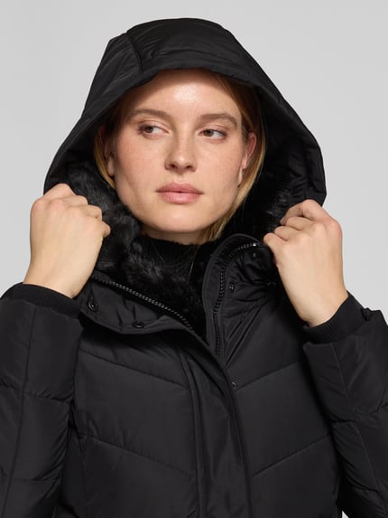 Frauenmodel trägt Steppmantel mit Kapuze von NAUTGÅRD 'NEBULA' in Schwarz, sichtbare Steppnähte, verdeckter Reißverschluss, Nahaufnahme Oberkörper. Regular Fit, Reißverschlusstaschen, Maße bei Größe S: Rückenlänge 108 cm, Schulterbreite 42 cm, Brustweite 37 cm, Ärmellänge 67 cm.