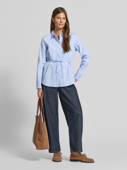 Frauenmodel trägt Regular Fit Hemdbluse aus reiner Baumwolle Modell 'ELINA' von VERO MODA, hellblau-weiß gestreift, kombiniert mit dunkelblauer, weiter Jeans und brauner Tasche, Farben in Blau, Weiß und Braun, glatte Texturen, Ganzkörperansicht von vorne, Damen Hemdbluse von VERO MODA.