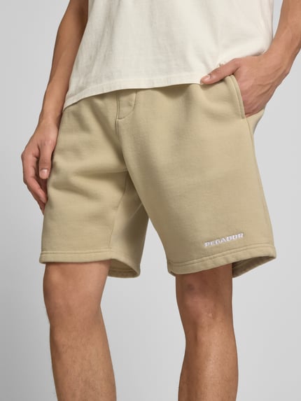 Männermodel trägt Shorts mit elastischem Bund und Logo-Stitching von Pegador, Beige, Baumwoll-Mix, Loose-Fit, Nahaufnahme Oberkörper. Bundweite 38 cm, Innenbeinlänge 23 cm, französische Eingrifftaschen.