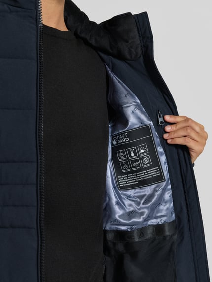 Frauenmodel trägt Jacke mit abnehmbarer Kapuze Modell 'ECLIPSE' von NAUTGÅRD in Dunkelblau mit silberfarbenem Futter und Label-Patch, Nahaufnahme Oberkörper. Kontext: Damen Jacke, gerader Schnitt, zweiwege-Reißverschluss, seitliche Eingrifftaschen mit Druckknopfverschluss, gestrickte Ärmelabschlüsse, Rückenlänge Größe S 79 cm, Schulterbreite 38 cm, Brustweite 48 cm, Ärmellänge 62 cm.