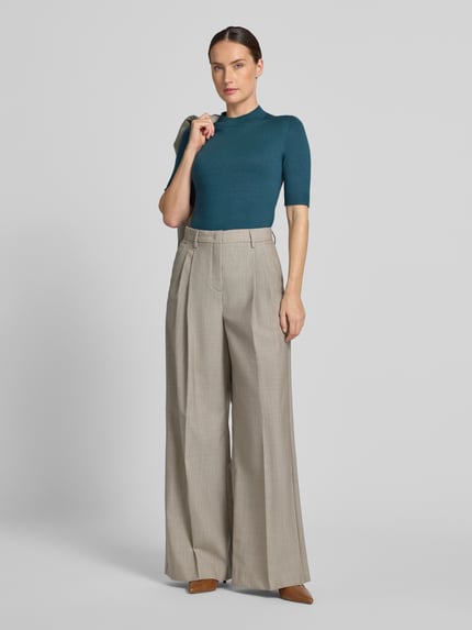 Frauenmodel trägt Slim Fit Strickpullover aus Viskose-Mix von COMMA in Dunkelgrün, kombiniert mit weiter beigefarbener Hose, Ganzkörperansicht von vorne. Gerippte Blenden und Abschlüsse, Halbarm, Rückenlänge 55 cm, Ärmellänge 29 cm.