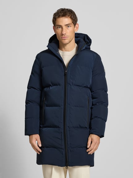 Männermodel trägt Mantel mit Kapuze und Label-Patch von NAUTGÅRD in Dunkelblau, gesteppte Optik, Zweiwege-Reißverschluss, Ganzkörperansicht von vorne. Rückenlänge 100 cm, Schulterbreite 52 cm, Brustweite 60 cm, Ärmellänge 65 cm.