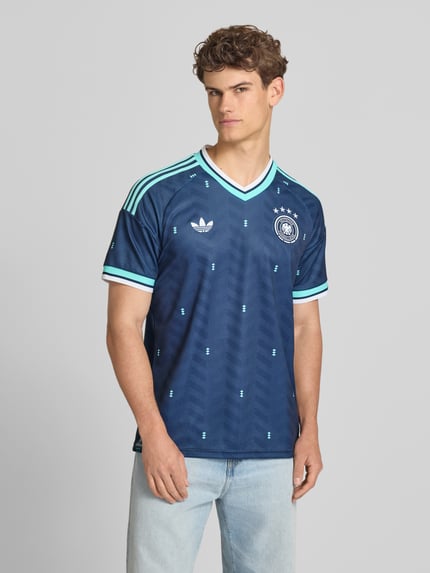 Męski model nosi DFB Auswärtstrikot Deutschland 2026 od adidas Sportswear, efekt: wielokolorowy, krój slim fit, aplikacja z logo, dekolt w serek, krótki rękaw, całkowity widok z przodu.