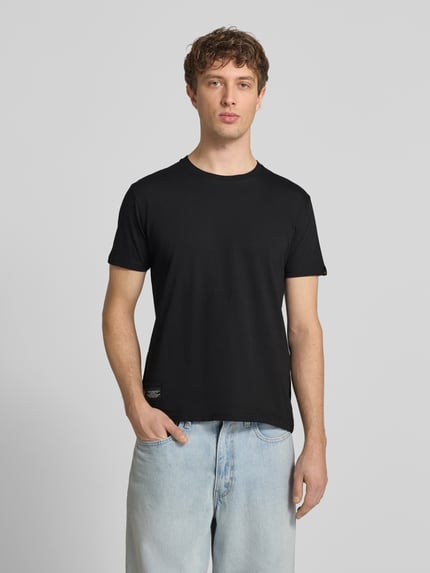 Männermodel trägt T-Shirt mit Label-Patch von Alpha Industries in Schwarz, Reine Baumwolle, Gerader-Schnitt, Rundhalsausschnitt, Ganzkörperansicht von vorne. Rückenlänge bei Größe M: 66 cm.