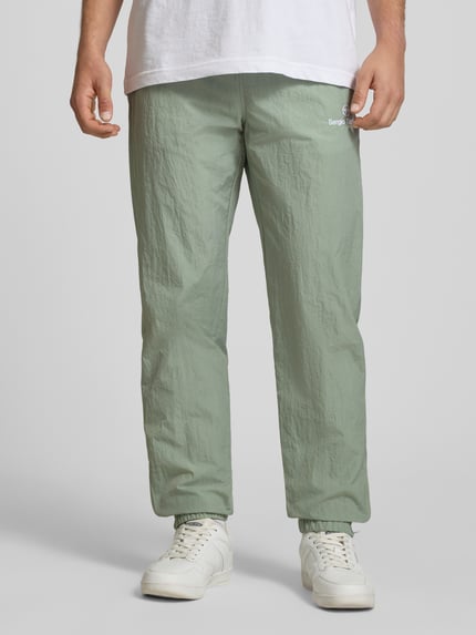 Männermodel trägt Straight Fit Stoffhose mit Logo-Stitching Modell 'Nayla' von SERGIO TACCHINI in Hellgrün, leichte Struktur, elastischer Bund mit Tunnelzug, elastische Beinabschlüsse, Innenbeinlänge 77 cm bei Größe M, Ganzkörperansicht von vorne.