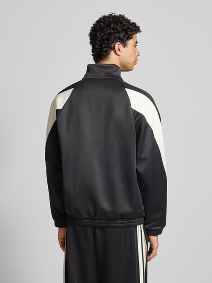 Męski model nosi Kurtkę treningową z logo i stójką od adidas Sportswear, mieszanka materiałów z bawełną, krój regular fit, długość tyłu 68 cm, długość rękawa 67 cm, kolor czarno-biały, widok całej sylwetki z tyłu.