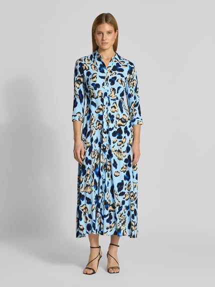 Frauenmodel trägt Hemdblusenkleid aus Viskose Modell 'SAVANNA' von YAS mit blauem, schwarzem und beigefarbenem Muster, Ganzkörperansicht von vorne. Taillierte Passform, Midilänge, Kentkragen, Knopfleiste, 3/4-Arm, Rückenlänge 135 cm, Brustweite 42 cm, Ärmellänge 49 cm.