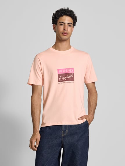Męski model nosi t-shirt z nadrukiem z napisem, woskowana bawełna, krój regular fit, okrągły dekolt, model 'RHYERES' od Jack & Jones, długość tyłu w rozmiarze M: 67 cm, jasnoróżowy kolor, gładka tekstura, widok całej sylwetki od przodu.