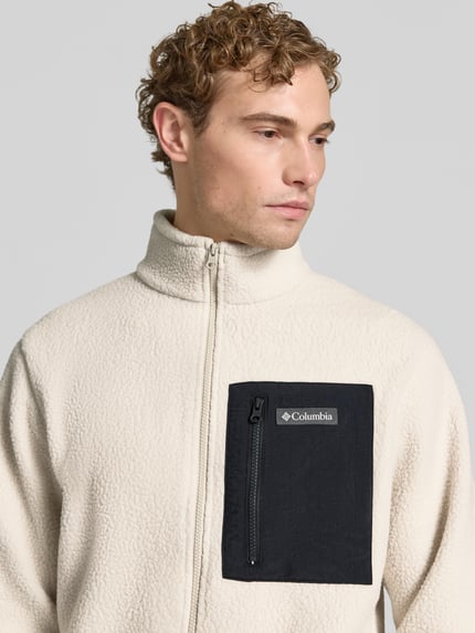 Männermodel trägt Fleecejacke mit Stehkragen Modell 'Jasper' von Columbia in Beige mit schwarzer Brusttasche, Nahaufnahme Oberkörper. Gerade Passform, Label-Patch, durchgehender Reißverschluss, Brusttasche mit Reißverschluss, seitliche Eingrifftaschen, elastische Ärmelabschlüsse, strukturierte Fleece-Textur.