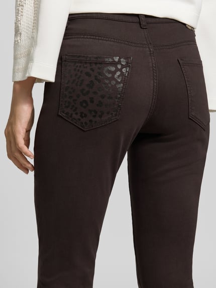 Kobieta nosi Slim Fit Jeans mit Lyocell-Anteil od Marc Cain, elastyczna mieszanka materiałów, 5 kieszeni, Długość 7/8, brąz, Szerokość w rozmiarze 36: 36 cm, Długość wewnętrzna nogawki: 72 cm, detal kieszeni z nadrukiem, zapięcie na guzik i zamek błyskawiczny, zdjęcie z tyłu, zbliżenie na biodra.
