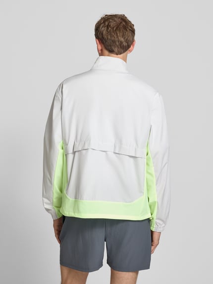 Männermodel trägt Regular Fit Jacke in Stehkragen Modell 'Fast Days' von New Balance, Farbkontrast Weiß und Neon-Grün, elastische Ärmelabschlüsse, Tunnelzug im Bund, Rückenlänge 68 cm, Eingrifftaschen mit Reißverschluss, Ganzkörperansicht Rückansicht.