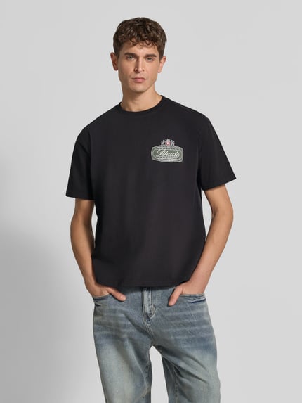 Männermodel trägt T-Shirt mit Label-Print von RHUDE aus reiner Baumwolle, locker geschnitten, dunkel, Rundhalsausschnitt, Ganzkörperansicht von vorne.