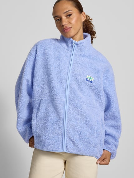 Frauenmodel trägt Regular Fit Jacke aus Teddyfell Modell 'Hoktown' von American Vintage, hellblau, Stehkragen, Logo-Patch, Reißverschluss, Eingrifftaschen, grob strukturierte Oberfläche, Ganzkörperansicht von vorne.
