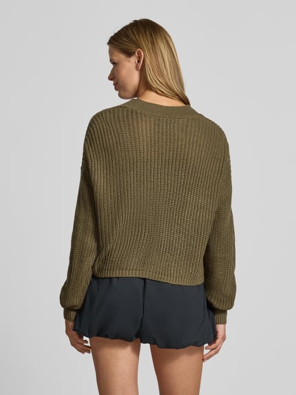 Frauenmodel trägt Boxy Fit Strickjacke mit V-Ausschnitt Modell 'CAROL' von ONLY in Olivgrün, grob gestrickt, Damen Strickjacke, Ganzkörperansicht Rückansicht.