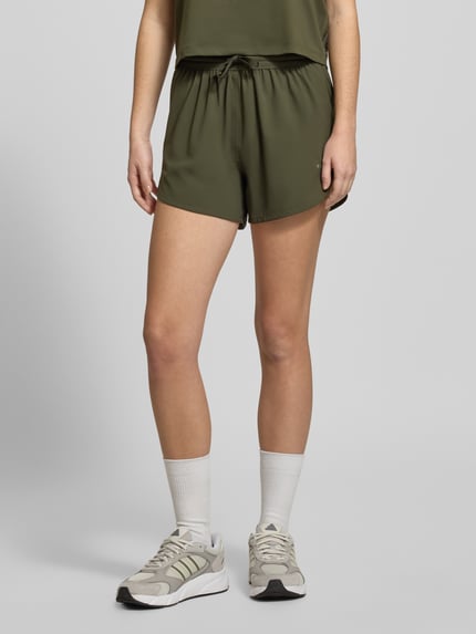 Vrouwenmodel draagt PUMA x HYROX - Shorts met labelprint in olijfgroen, los model, elastische band met tunnelkoord, binnenbeenlengte 11 cm, bandwijdte 36 cm, labelprint-detail, voorkant bovenbeen, close-upvoorname.
