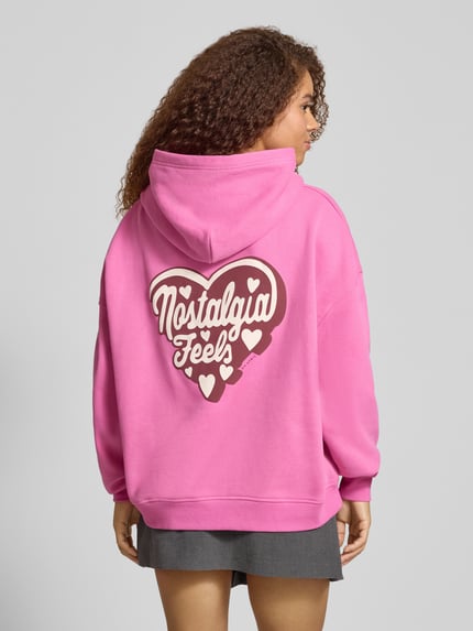 Frauenmodel trägt Hoodie mit rückseitigem Motiv-Print Modell 'Nostalgia Heart' von OH APRIL, pink, oversized, Baumwoll-Mix, Känguru-Tasche, Ganzkörperansicht Rückansicht.