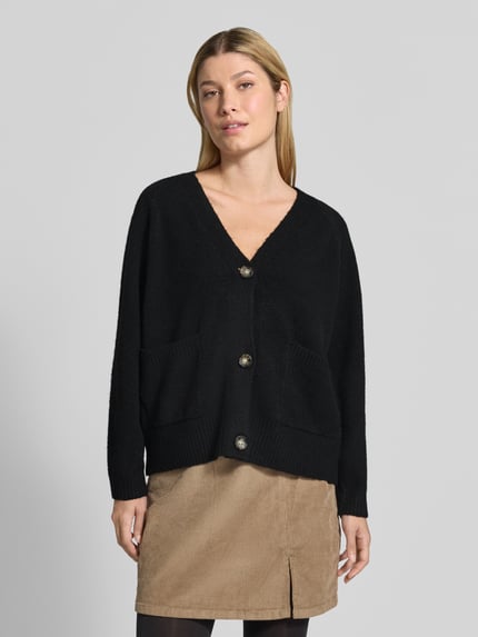 Frauenmodel trägt Oversized Strickjacke mit Woll-Anteil Modell 'BOOM' von VERO MODA in Schwarz, beige Rock, glatte Strickstruktur, Ganzkörperansicht von vorne.