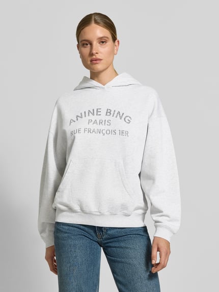 Frauenmodel trägt Oversized Hoodie aus reiner Baumwolle von Anine Bing in Hellgrau mit Label-Print, Känguru-Tasche, elastischen Abschlüssen, Ganzkörperansicht von vorne.