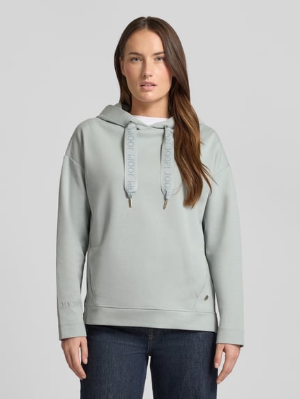 Frauenmodel trägt Regular Fit Hoodie mit Logo-Applikation von JOOP!, Baumwoll-Mix, Kapuze mit Tunnelzug, elastische Abschlüsse, Eingrifftaschen, verlängerte Rückseite, hellgrau, glatte Textur, Ganzkörperansicht von vorne.