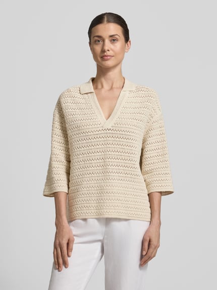 Modelka kobieca ma na sobie sweter z dzianiny z dekoltem w serek i rękawem o dł. 3/4 model 'Garakino' od OPUS, z czystej bawełny, krój relaxed fit, kołnierzyk polo, długość tyłu w rozmiarze 36: 56 cm, długość rękawów w rozmiarze 36: 33 cm, kremowy kolor, widoczne ażurowe faktury, całkowity widok od przodu.