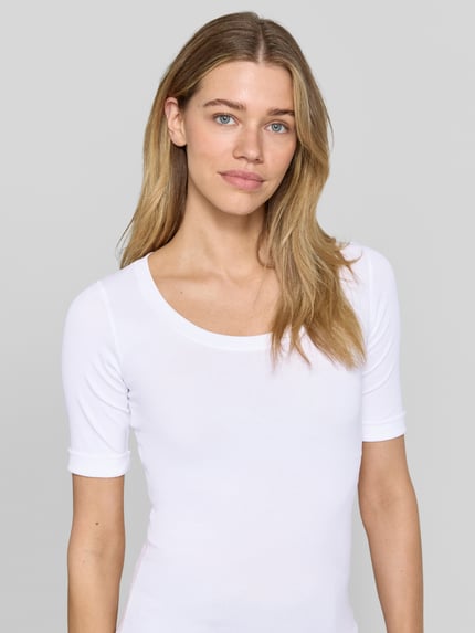 Modelka kobieca ma na sobie T-Shirt mit Ärmelaufschlag und Rundhalsausschnitt od Marc Cain, mieszanka bawełny z dodatkiem streczu, dopasowany krój, okrągły dekolt, rękawy z zakładką, detal z logo, długość tyłu w rozmiarze 36: 60 cm, biały kolor, gładka faktura; zbliżenie na górną część ciała z przodu.
