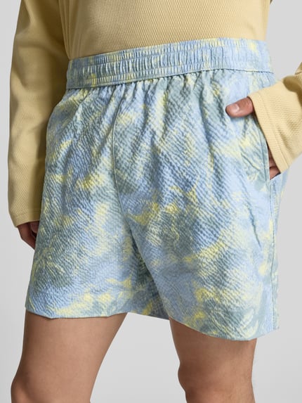 Męski model nosi spodenki kąpielowe z fakturowanym wzorem od MAISON KITSUNÉ, prosty krój, elastyczny pas, wpuszczane kieszenie, kieszenie z tyłu, długość wewnętrzna nogawki w rozmiarze M 13 cm, szerokość w rozmiarze M 42 cm, pastelowe kolory, całkowity widok z przodu.