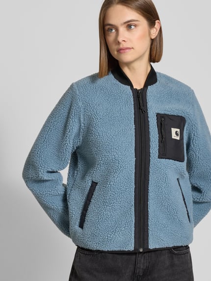Frauenmodel trägt Fleecejacke im Allover-Look mit Reißverschluss Modell 'Janet' von Carhartt Work In Progress in Hellblau und Schwarz, Label-Detail, Brusttasche, Stehkragen, Regular Fit, Nahaufnahme Oberkörper.