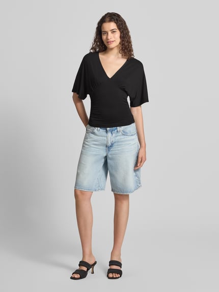 Vrouwenmodel draagt Bluse aus Viskose-Mix mit V-Ausschnitt van Gina Tricot in zwart, getailleerde pasvorm, halflange mouwen, Ruglengte maat S 26 cm, Mouwlengte maat S 30 cm, Ganzkörperansicht van voren.