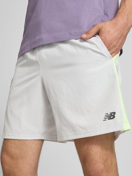 Mężczyznamodel nosi Shorts mit elastischem Bund und Logo od New Balance w kolorze białym z limonkowym akcentem, krój regular fit, logo, elastyczny pas z troczkiem, szerokość w rozmiarze M: 42 cm, długość nogawki od wewnątrz w rozmiarze M: 19 cm, zbliżenie na górną część nóg z przodu.
