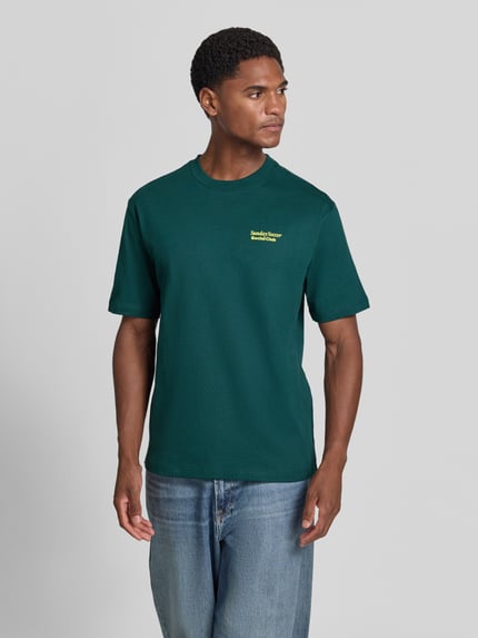Männermodel trägt T-Shirt mit Statement-Print von MCNEAL in Dunkelgrün mit gelbem Schriftzug, kombiniert mit Jeans, Ganzkörperansicht von vorne, lockerer Schnitt, Rundhalsausschnitt, Rückenlänge bei Größe M 66 cm.